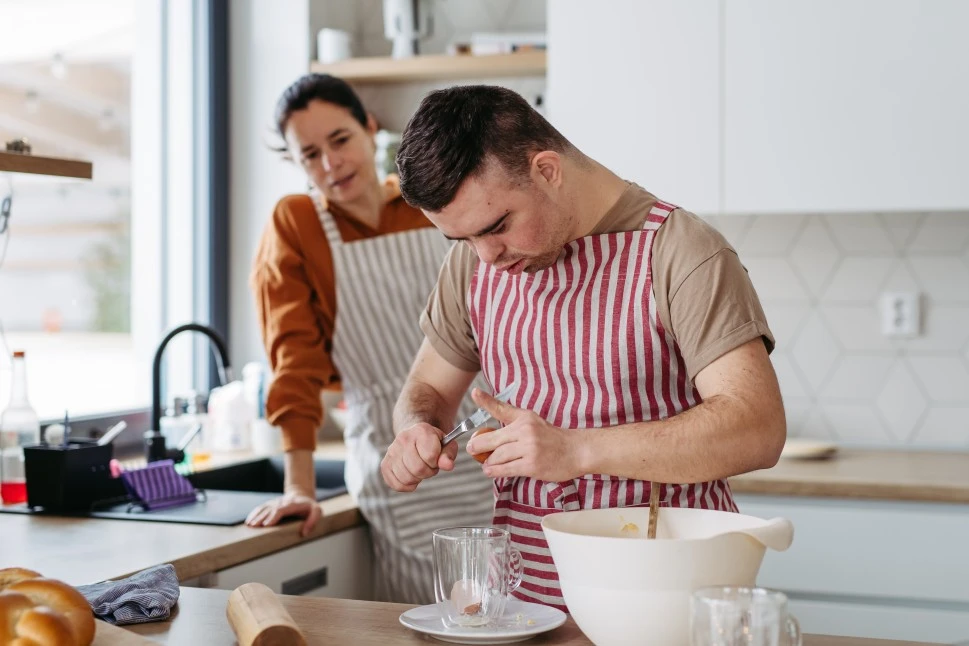 Support worker helping disabled man with down syndrome bake. Disabled man cracking egg into cup