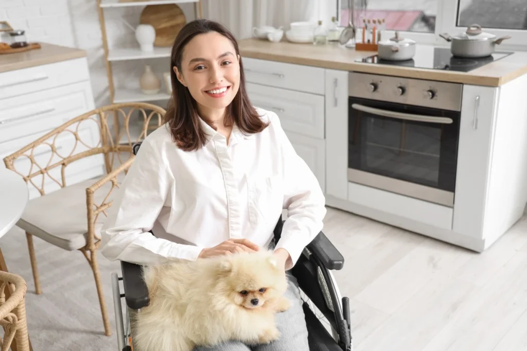 Disabled woman in wheelchair sitting in kitchen smiling at camera with dog on lap